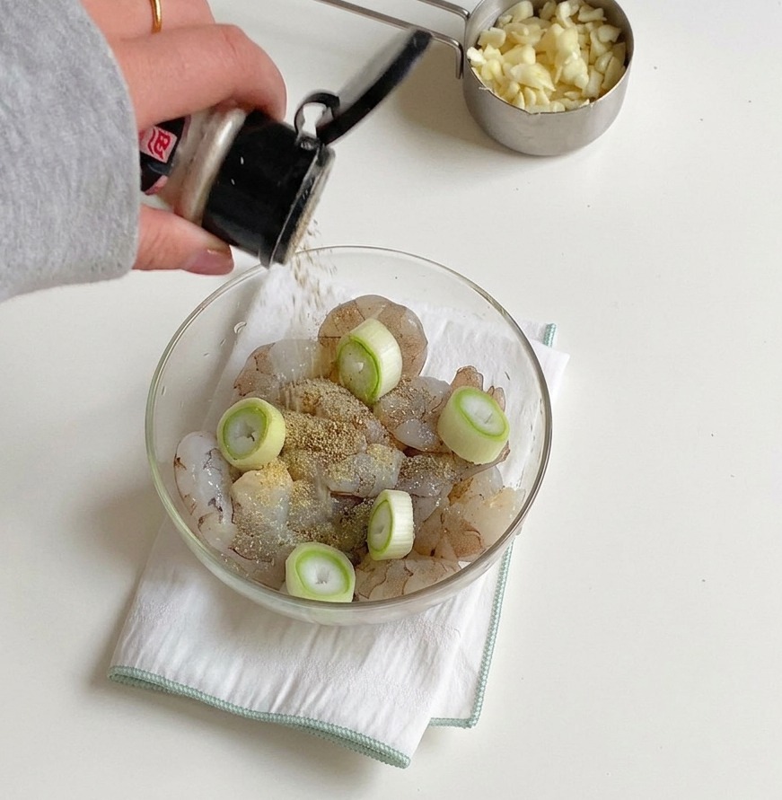 A hand shaking a seasoning container to add pepper and salt to a bowl of raw shrimp and green onion slices.