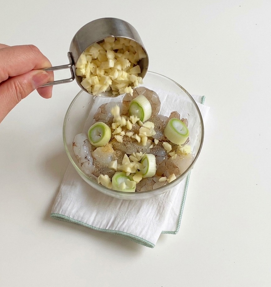 Minced garlic being poured from a small metal measuring cup into a bowl containing seasoned raw shrimp.