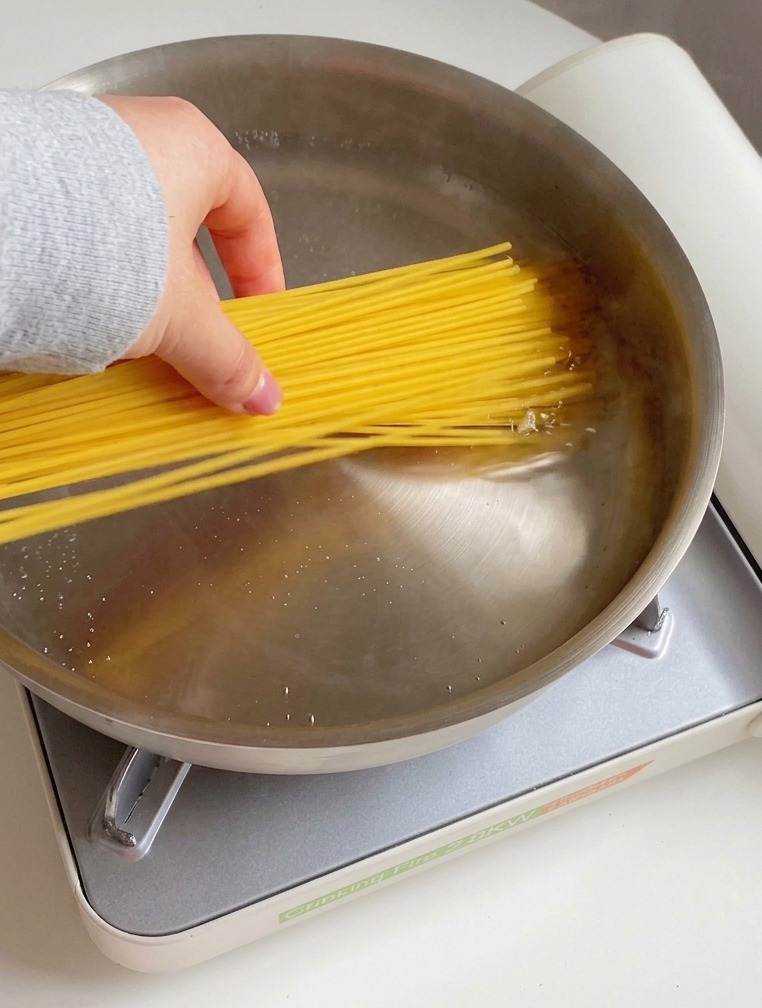 A hand placing a bundle of dry spaghetti into a stainless steel pan filled with water, oil droplets, and salt.