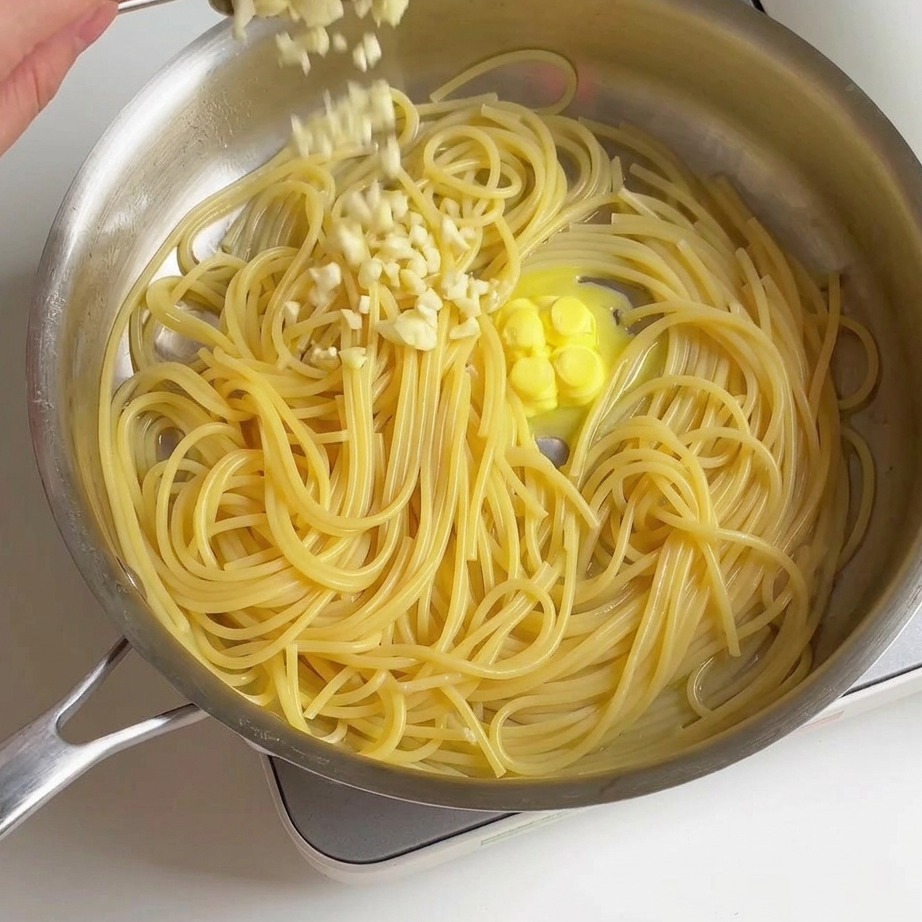 Freshly minced garlic being sprinkled from a spoon over cooked spaghetti and melting butter in a pan.