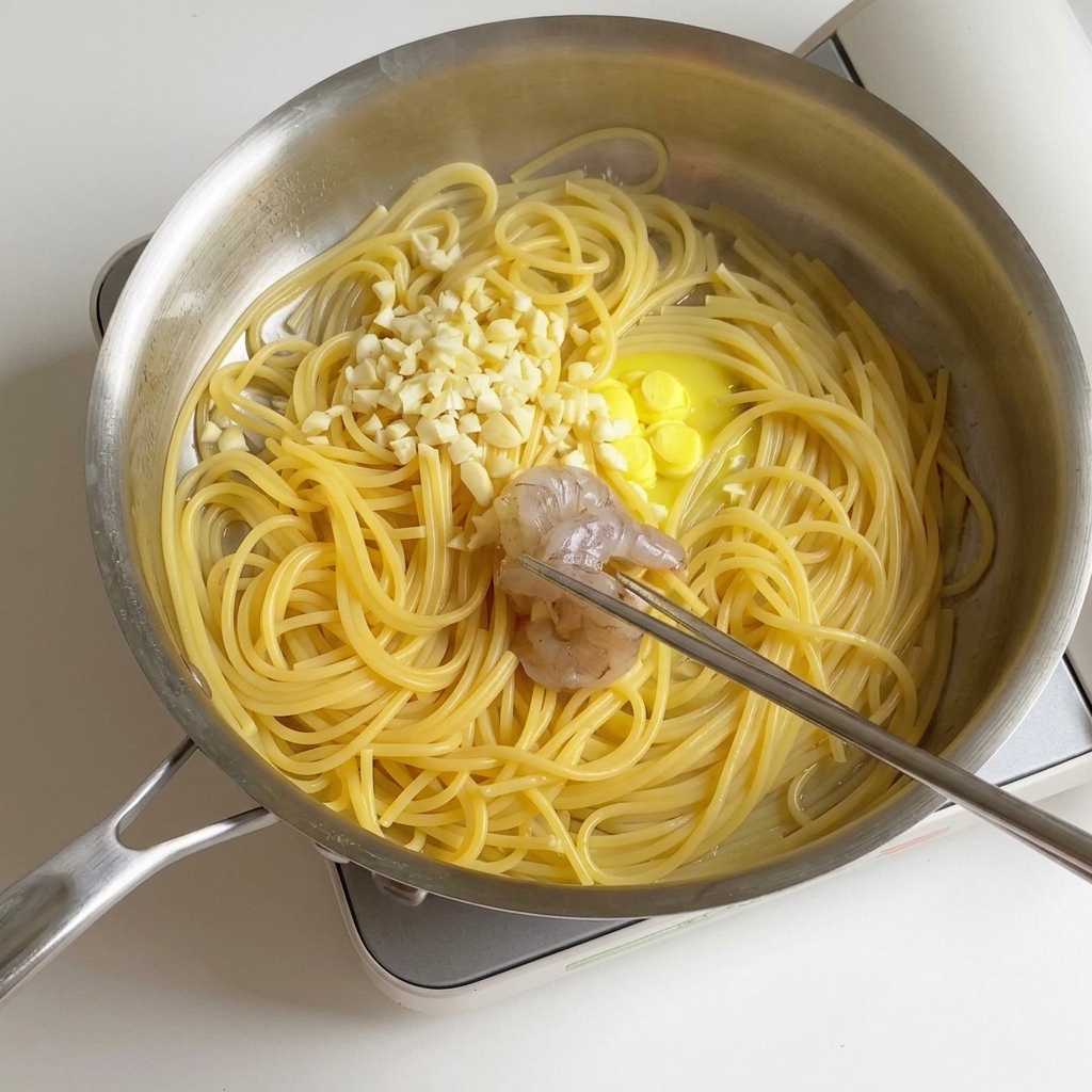 Marinated shrimp being placed into a stainless steel pan containing cooked spaghetti, melting butter, and minced garlic.