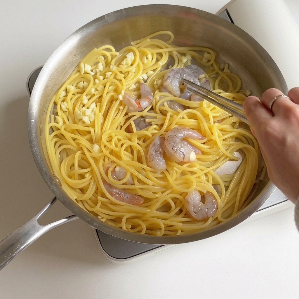 A person stir-frying shrimp and spaghetti in a pan as the shrimp turn pink and opaque.