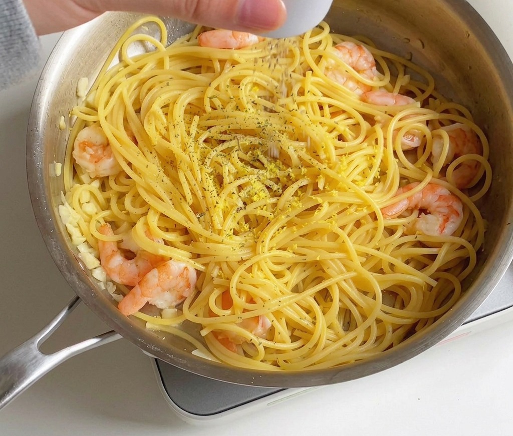 A hand seasoning a pan of shrimp and lemon spaghetti with dried parsley flakes and salt.