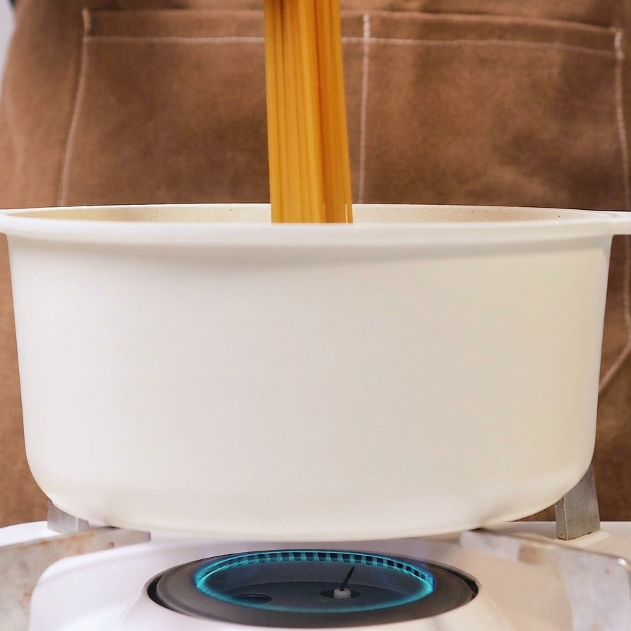 A person holding a bundle of dry spaghetti and placing it into a white pot of boiling water on a gas stove.