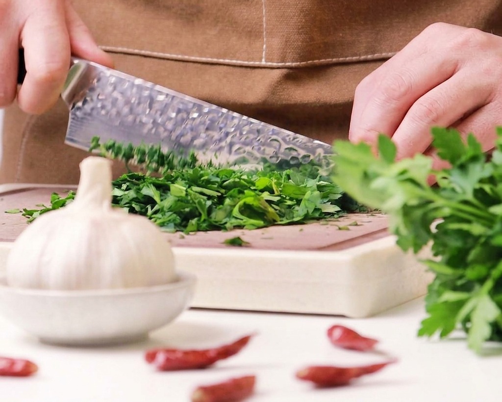 A close-up of a chef using a dimpled knife to finely mince a pile of fresh green parsley on a wooden board.