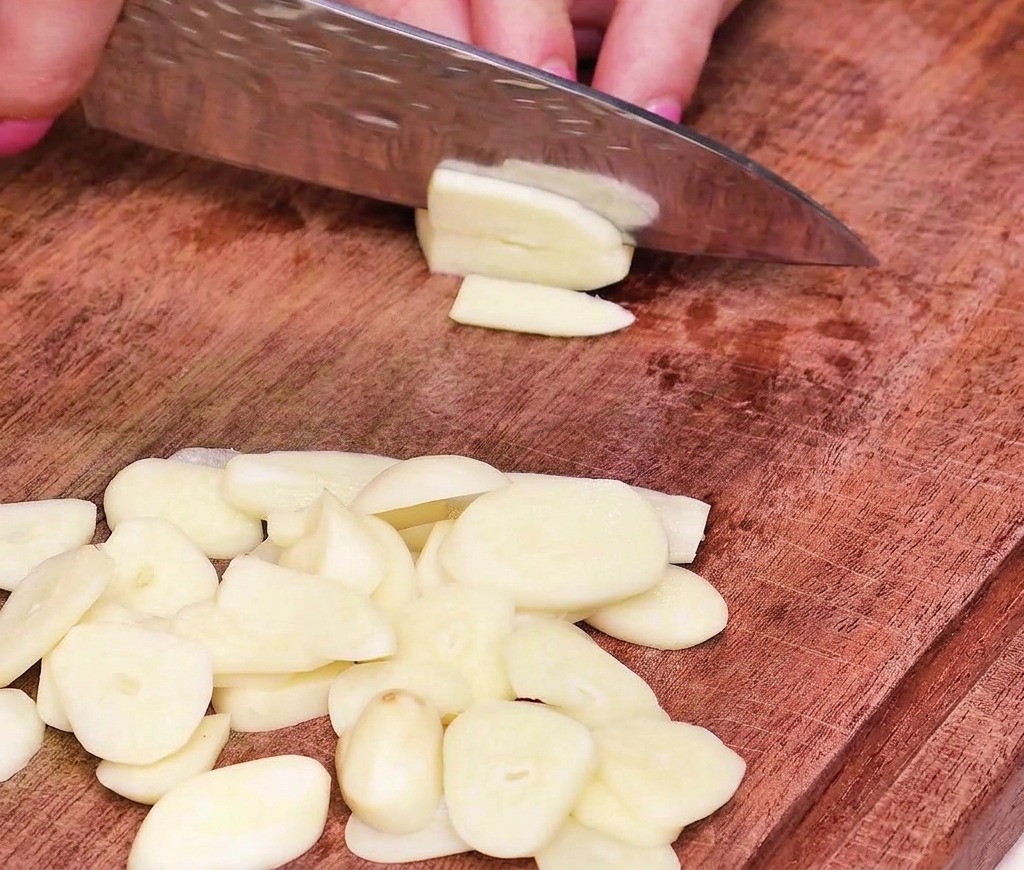 A close-up view of a hand using a professional chefs knife to slice garlic cloves into thin, uniform rounds on a wooden cutting board.