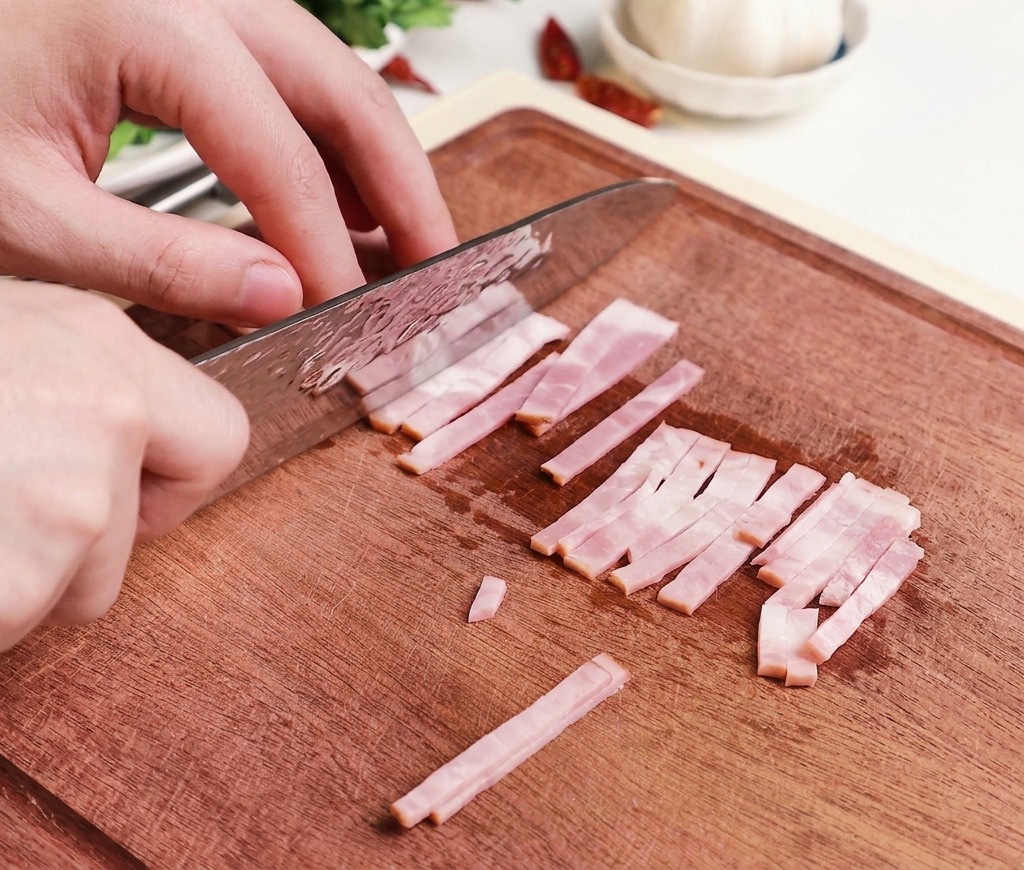 Hands using a professional chefs knife to slice raw pink bacon strips into small uniform pieces on a wooden cutting board.