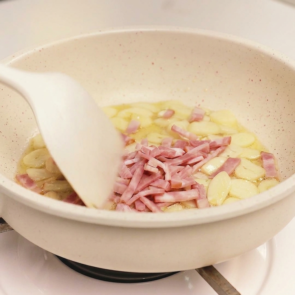 A white spatula stirring sliced bacon and garlic pieces together in a light-colored frying pan.