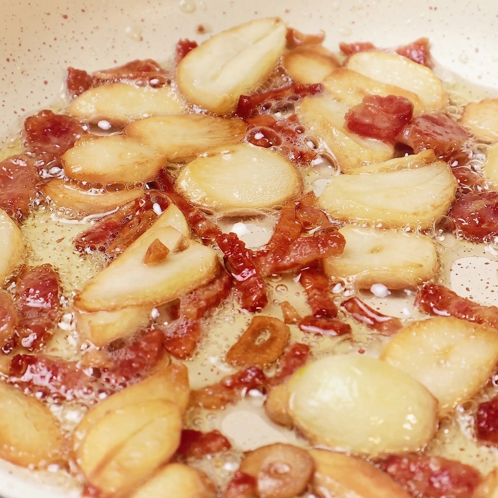 Garlic slices and small pieces of bacon bubbling in oil inside a pan, showing a rich golden-brown color and crispy texture.