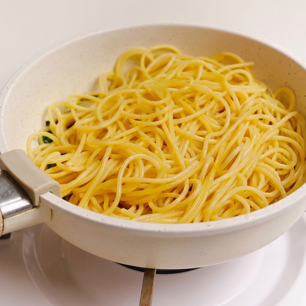 A batch of cooked spaghetti noodles sitting on top of the green herb, garlic, and bacon sauce in a light-colored pan.