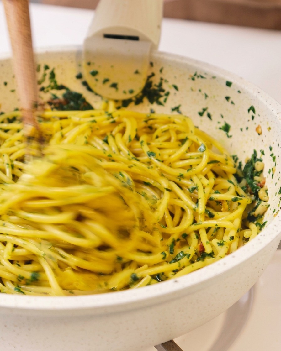 An action shot of spaghetti being tossed and mixed with green herbs and oil in a pan using a pair of wooden chopsticks.
