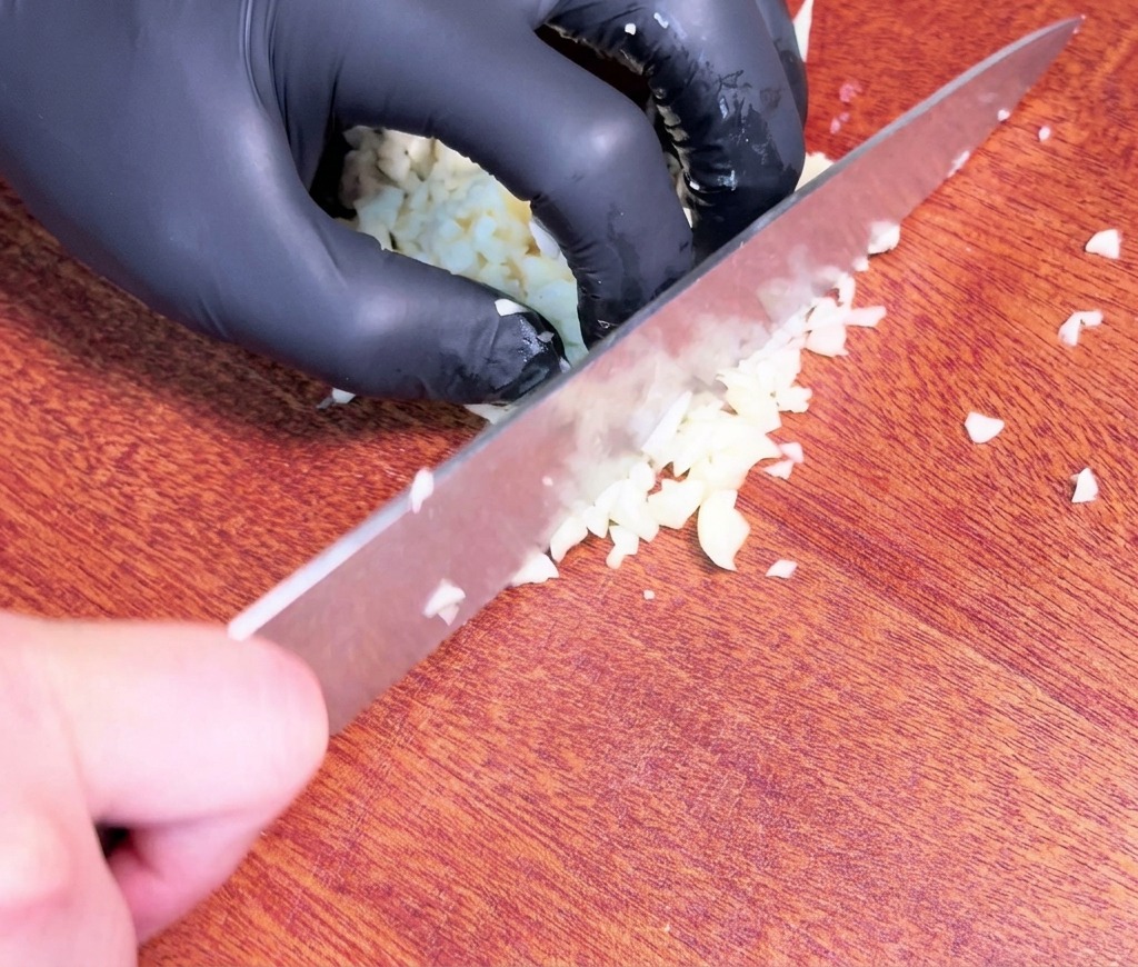 A hand wearing a black glove uses a large chefs knife to finely mince raw garlic cloves on a dark wooden cutting board.