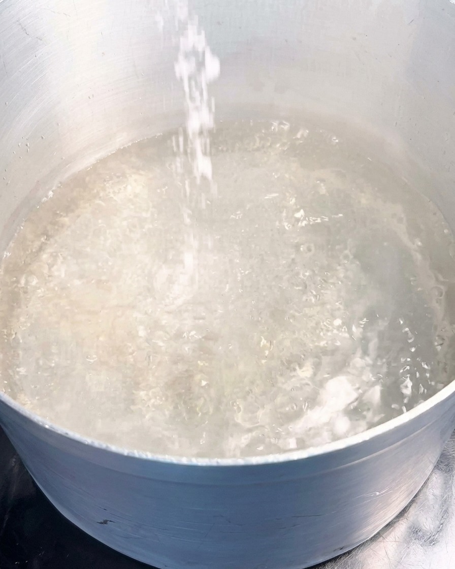 A stream of white sea salt being poured into a pot of vigorously bubbling boiling water.