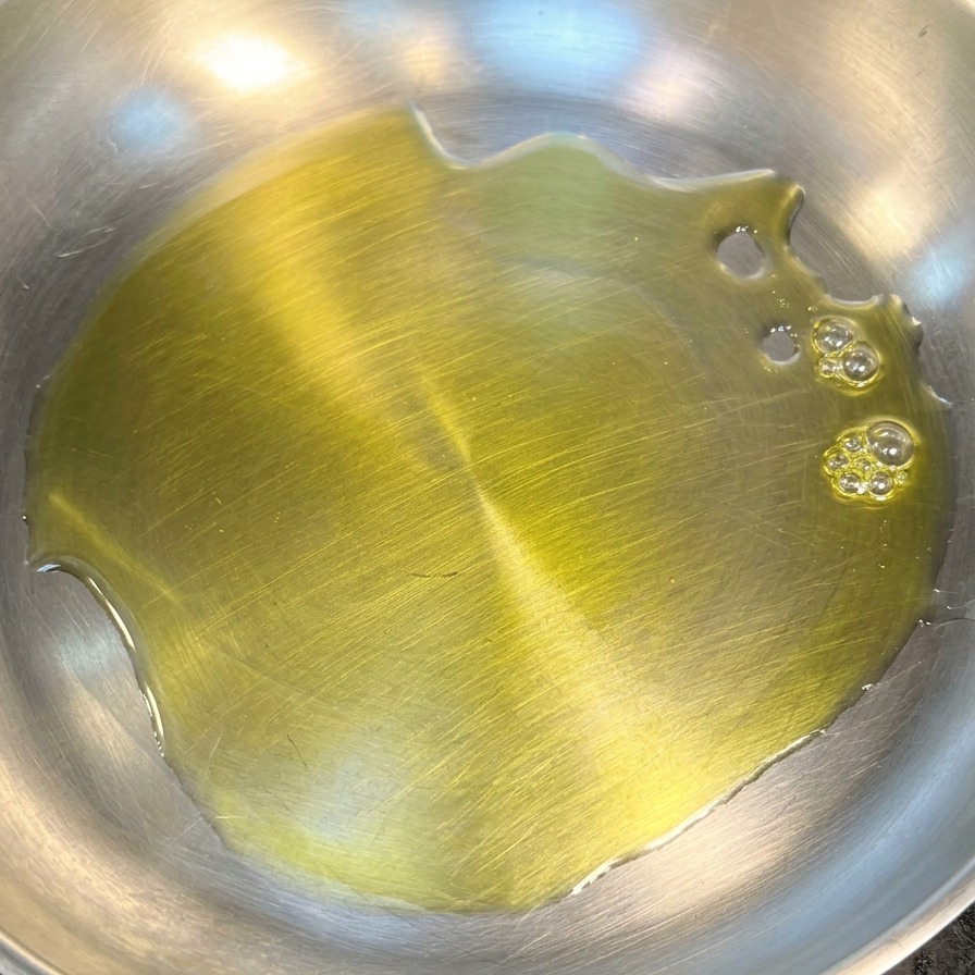 A pool of vibrant yellow olive oil coating the bottom of a clean stainless steel frying pan.