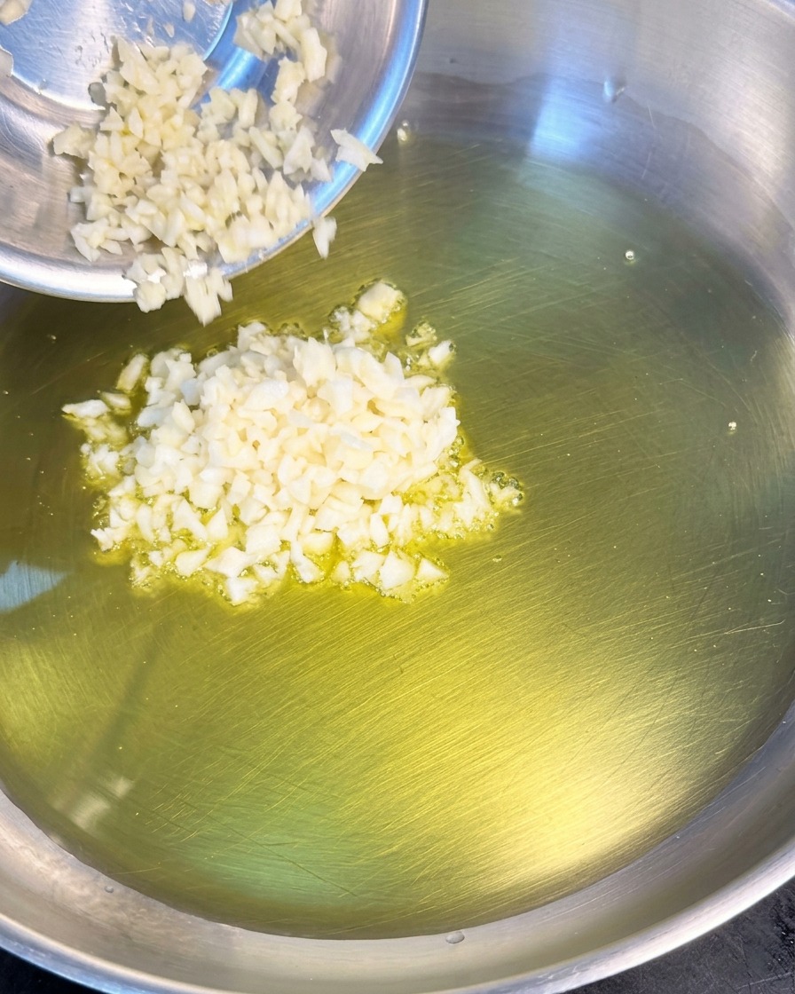 A pile of finely minced white garlic being added from a small metal bowl into a pan of shimmering olive oil.