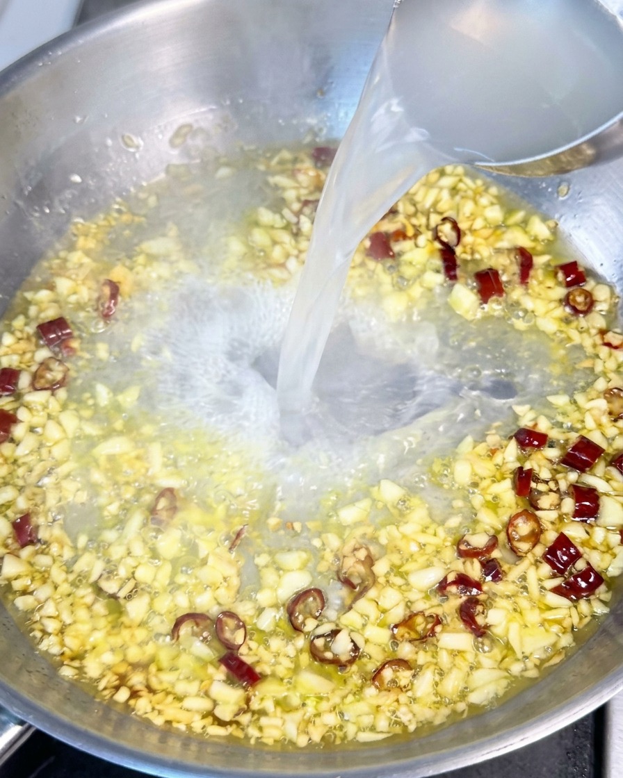 Cloudy white pasta water being poured from a ladle into a pan containing oil, garlic, and red chili flakes.