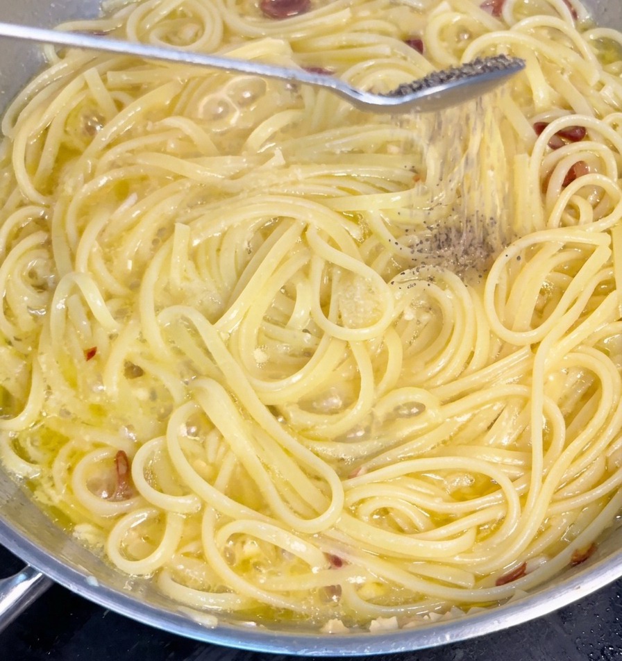 Freshly ground black pepper falling from a spoon onto spaghetti noodles simmering in a garlic and oil sauce.
