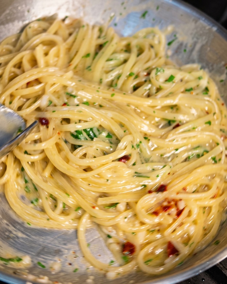 A stainless steel skillet filled with spaghetti being tossed with tongs. The noodles are coated in a glistening, creamy sauce with visible bits of minced garlic, red chili flakes, and fresh green parsley.