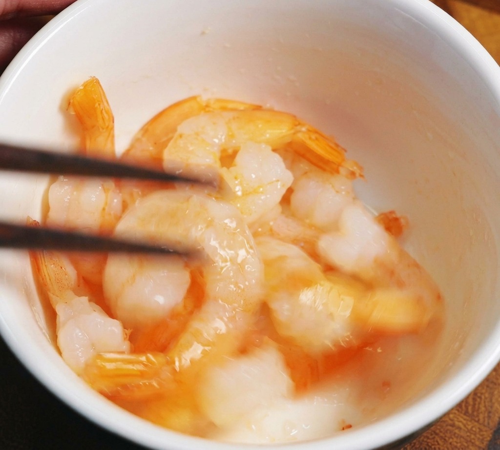 Raw, translucent shrimp being mixed with salt in a white bowl using wooden chopsticks.