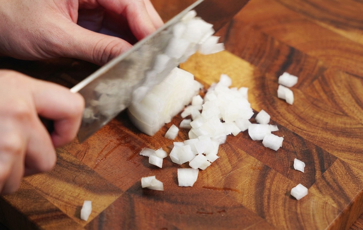 A persons hands using a chefs knife to dice a white onion on a wooden cutting board.