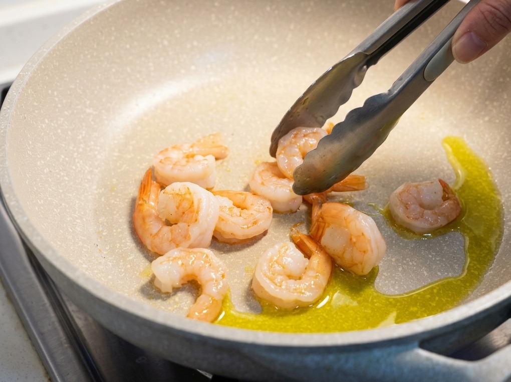 Cooked pink shrimp being turned with metal tongs in a light-colored pan with shimmering olive oil.