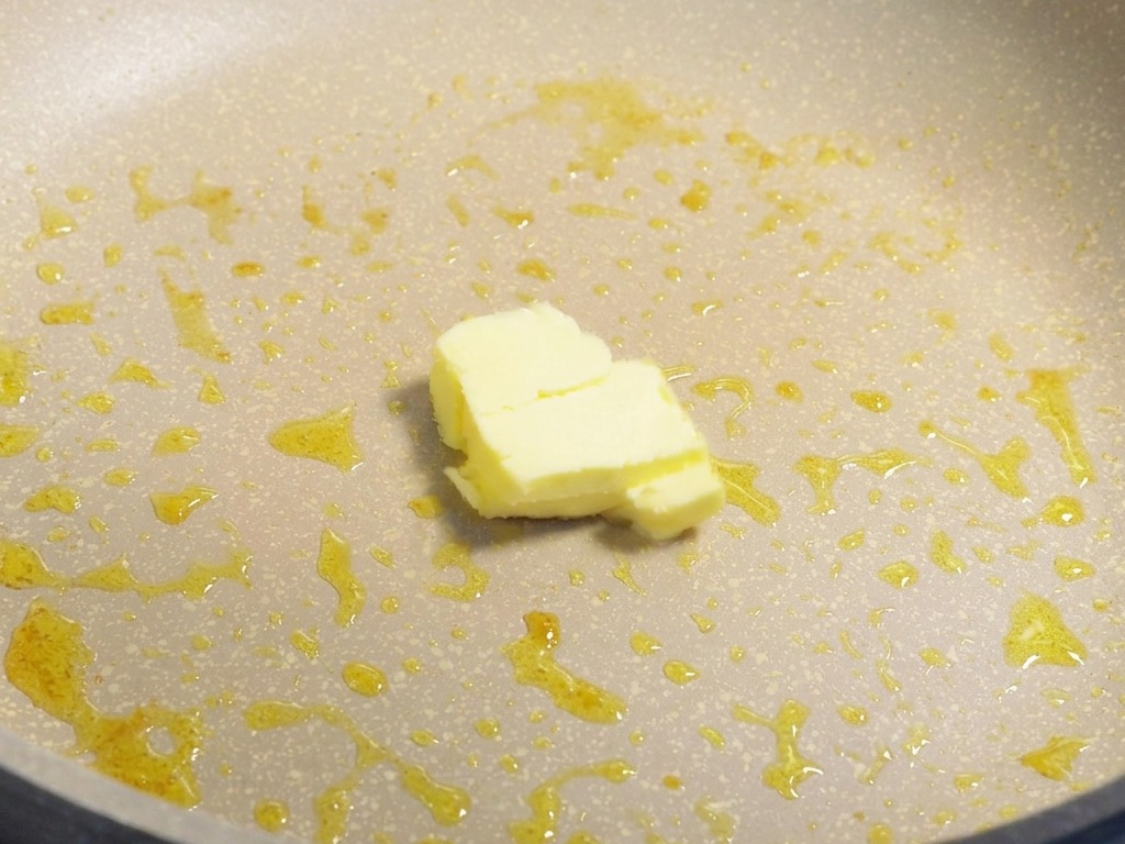 A small block of yellow butter melting in a pan containing residual cooking oil.