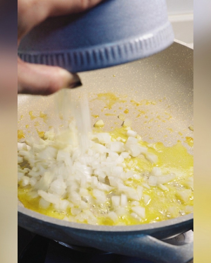 Diced white onions being added from a blue bowl into a pan of melted butter.