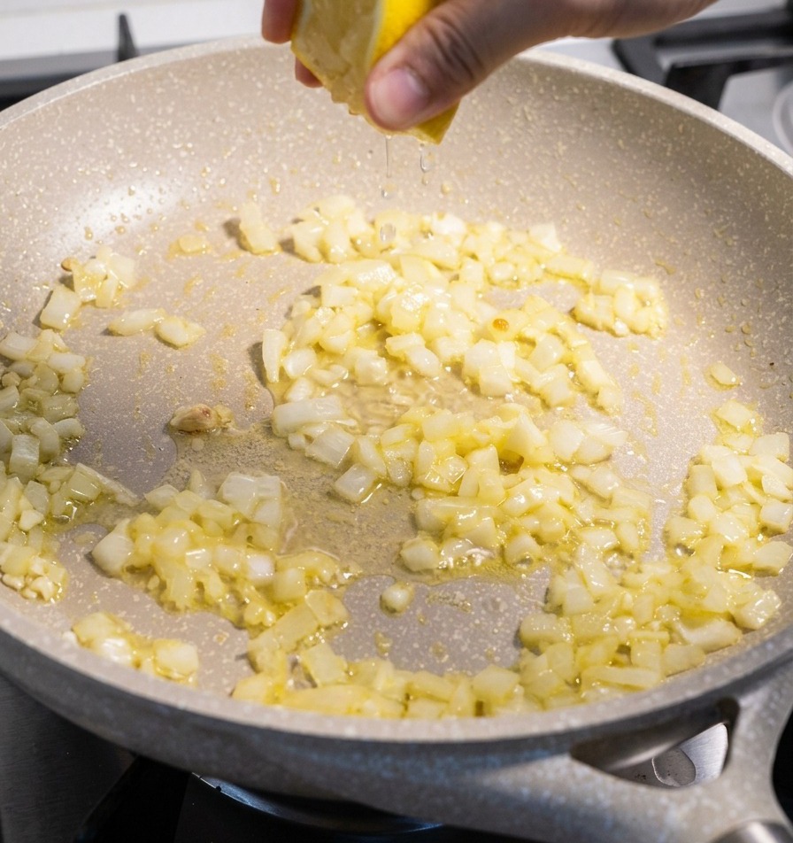 A hand squeezing a yellow lemon half over a skillet containing translucent sautéed onions and garlic pieces.