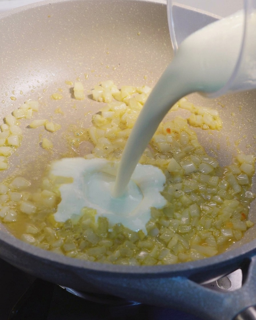 Heavy cream being poured from a measuring cup into a speckled beige pan containing sautéed onion and garlic bits.