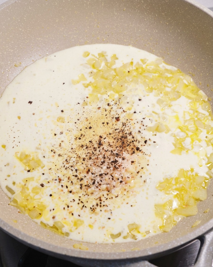 A pan of smooth white cream sauce with a pile of freshly ground black pepper and parmesan cheese in the center.
