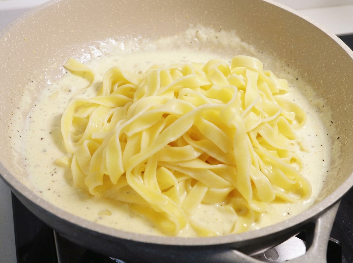 A large mound of cooked yellow fettuccine noodles resting on top of a thick, white bubbling cream sauce in a large pan.