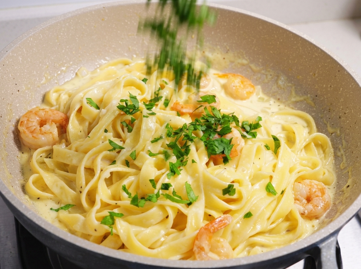 Chopped fresh green parsley being sprinkled over a creamy shrimp and fettuccine pasta dish in a light-colored pan.