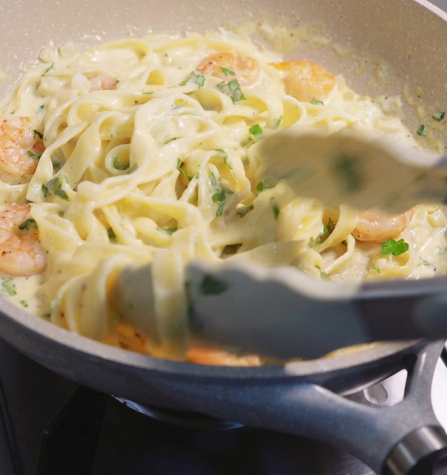 Using tongs to lift and mix fettuccine pasta and pink shrimp in a creamy white sauce within a speckled frying pan.