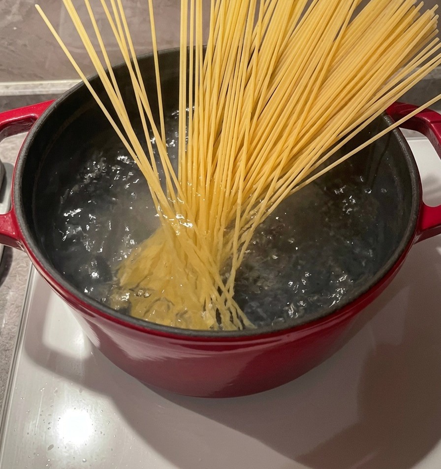 A bundle of dry spaghetti noodles being placed into a pot of bubbling boiling water using wooden chopsticks.