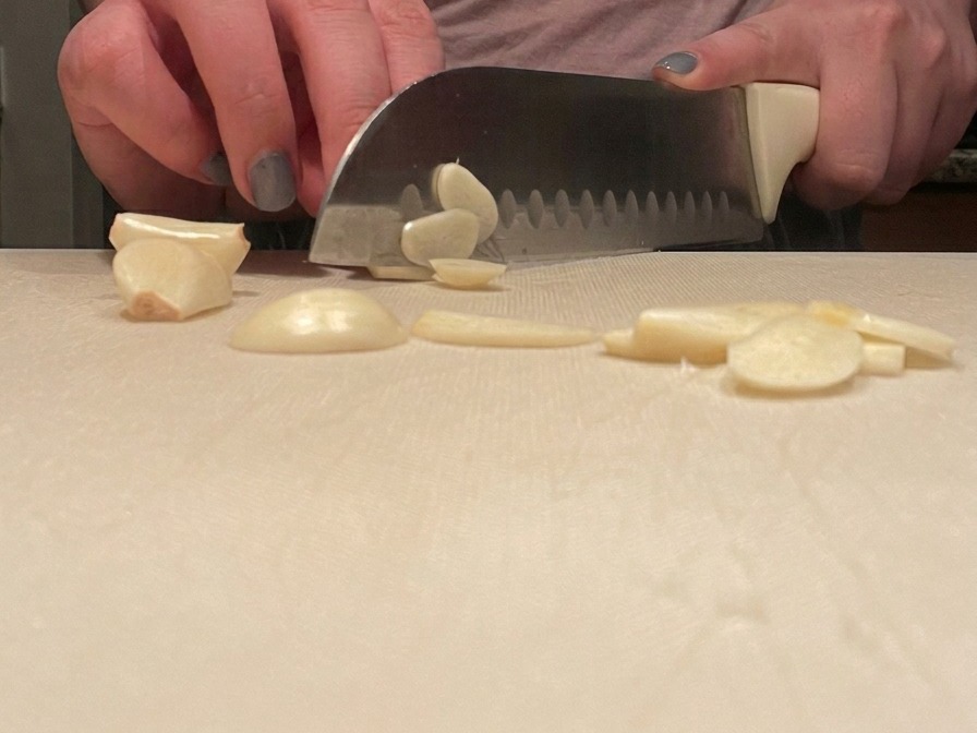 A persons hands carefully slicing garlic cloves into thin rounds with a chefs knife on a clean white cutting board.