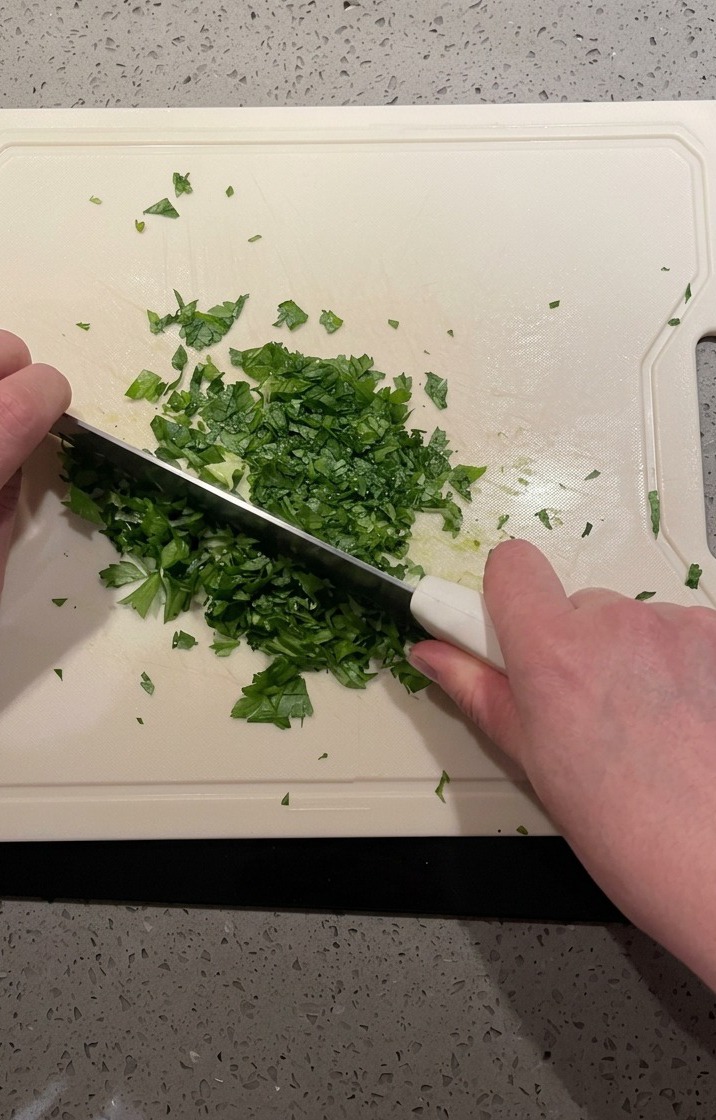 An overhead view of a knife finely mincing a pile of fresh green parsley on a white board.