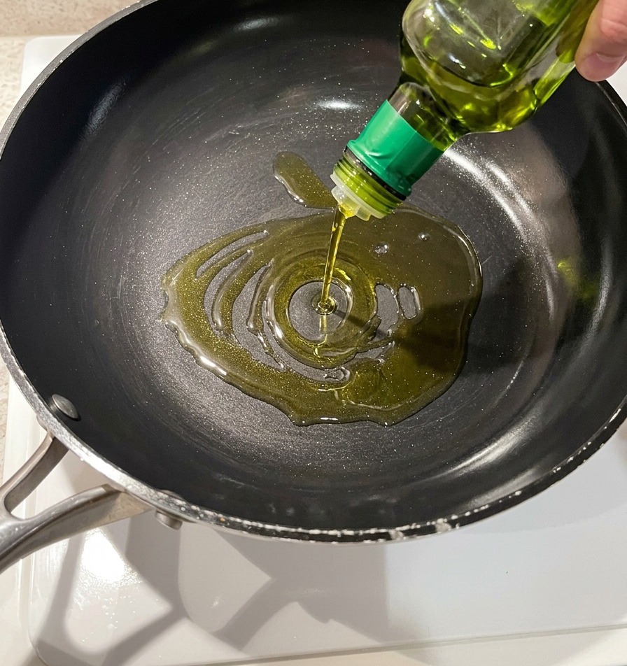 Golden olive oil being poured into a dark non-stick frying pan from a glass bottle.