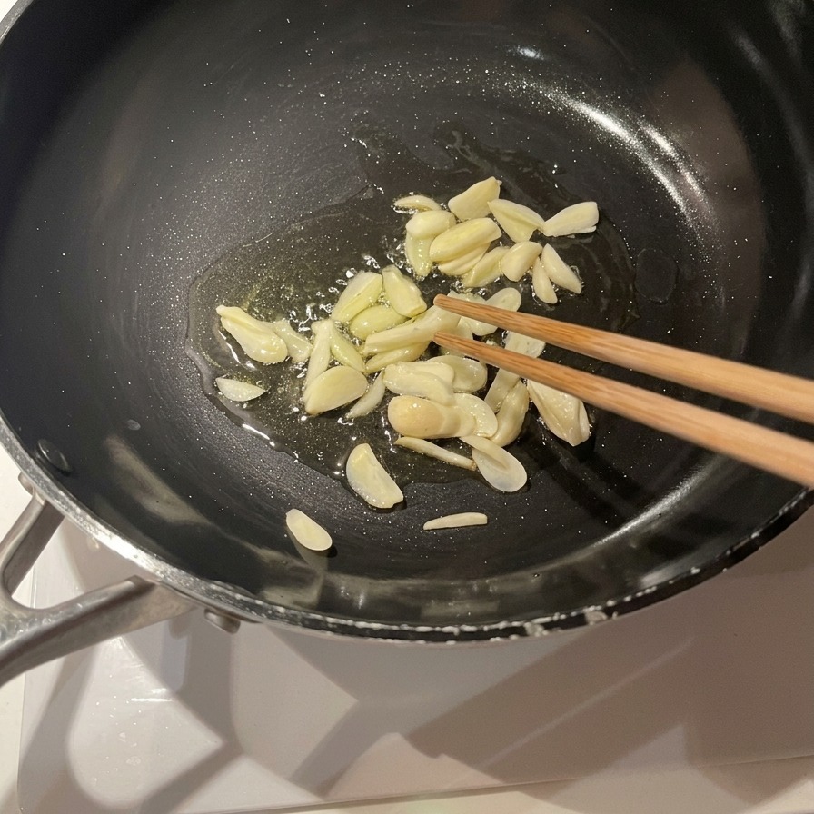 Sliced garlic cloves frying in a pool of olive oil in a black skillet, being stirred with wooden chopsticks.