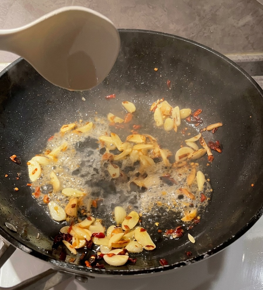 A white ladle pouring clear pasta water into a black pan containing sautéed garlic slices and red chili flakes.