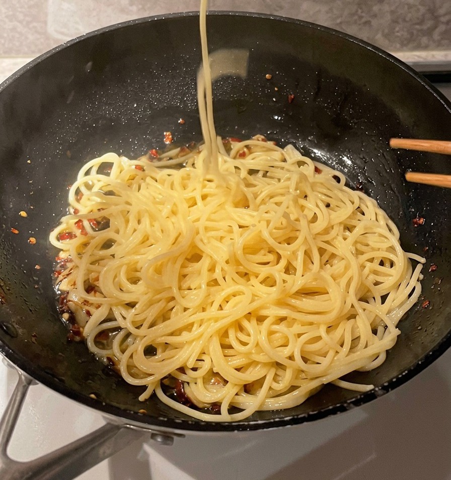 A metal mesh strainer dropping long strands of cooked spaghetti noodles into a pan containing garlic and oil.
