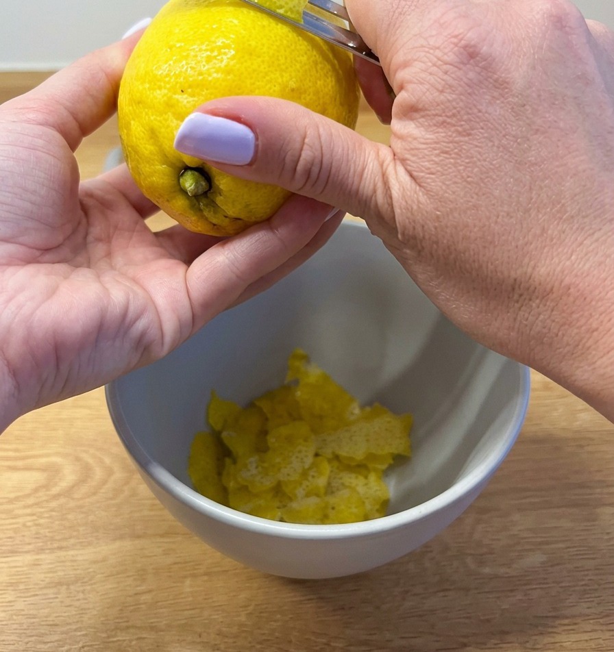 A close-up shot of hands using a metal vegetable peeler to remove long strips of bright yellow zest from a fresh lemon into a white bowl.