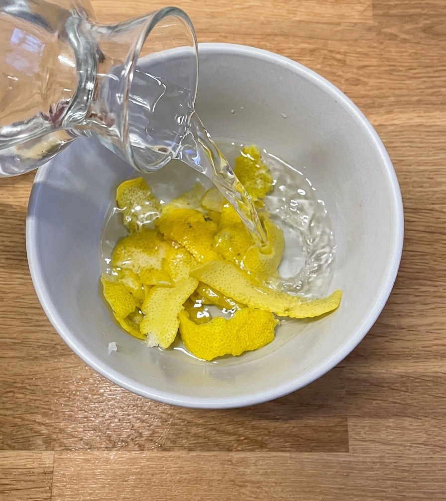 Clear water being poured from a glass carafe into a white ceramic bowl containing several large strips of fresh lemon peel.