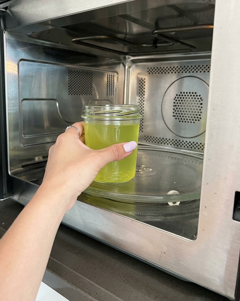 A hand placing a glass jar filled with yellow-tinted lemon water into a modern stainless steel microwave.