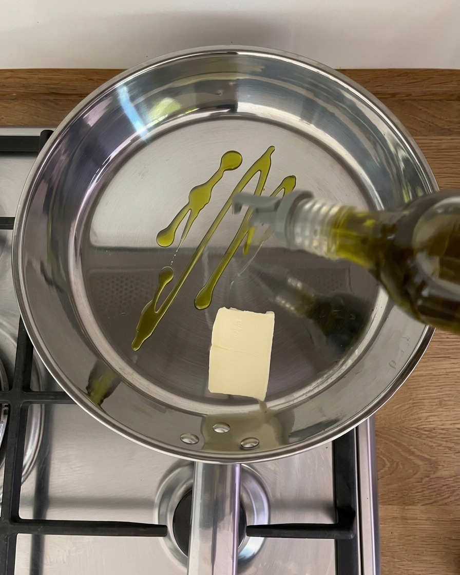 Olive oil being drizzled from a bottle into a shiny stainless steel frying pan containing a single pat of butter on a stovetop.