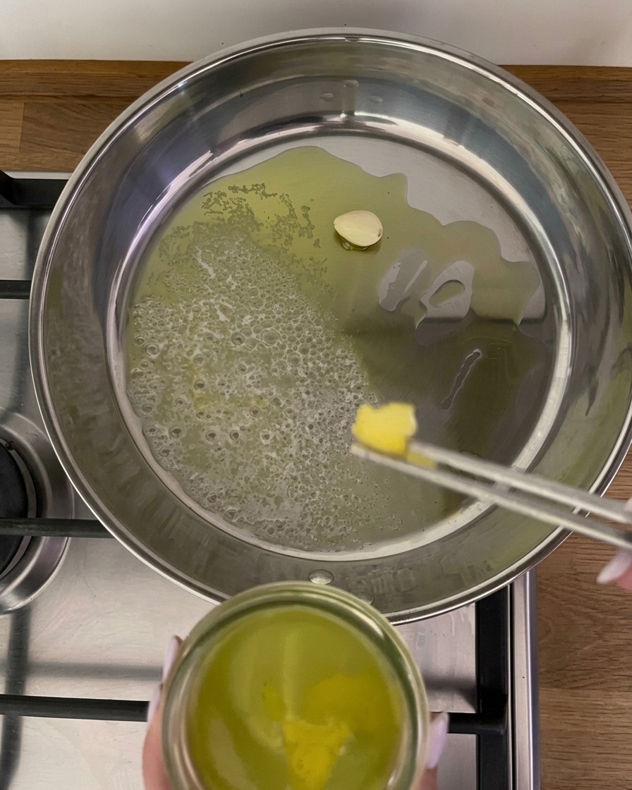 Metal tongs placing a strip of yellow lemon peel into a pan with bubbling butter, oil, and a single clove of garlic.