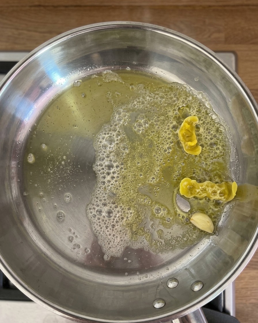 Warm lemon-infused water being poured into a stainless steel pan containing sautéed garlic and lemon peels, creating bubbles in the oil and butter mixture.