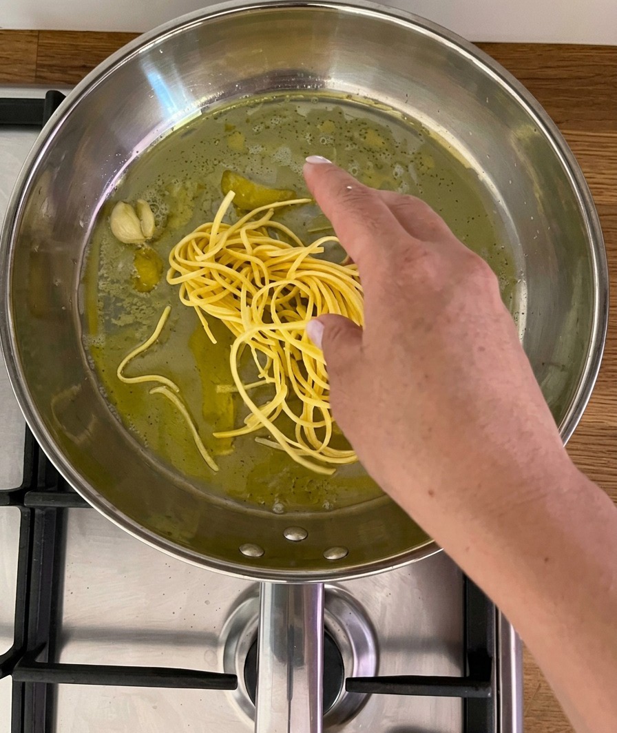 A hand placing a bundle of fresh yellow spaghetti-like noodles into a stainless steel pan containing oil, a garlic clove, and lemon peel.