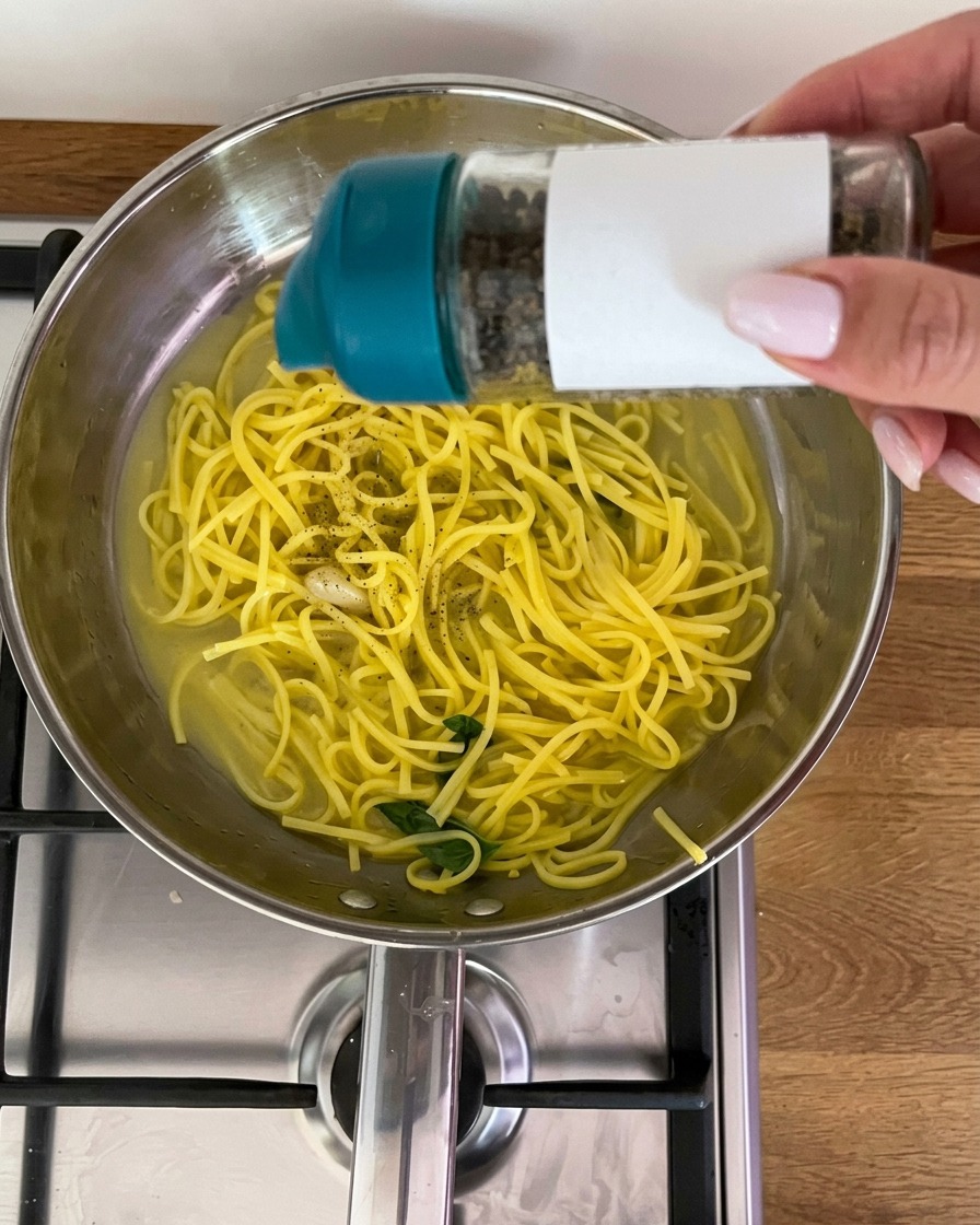 Freshly ground black pepper being shaken from a grinder over a pan of lemon pasta with fresh basil leaves.