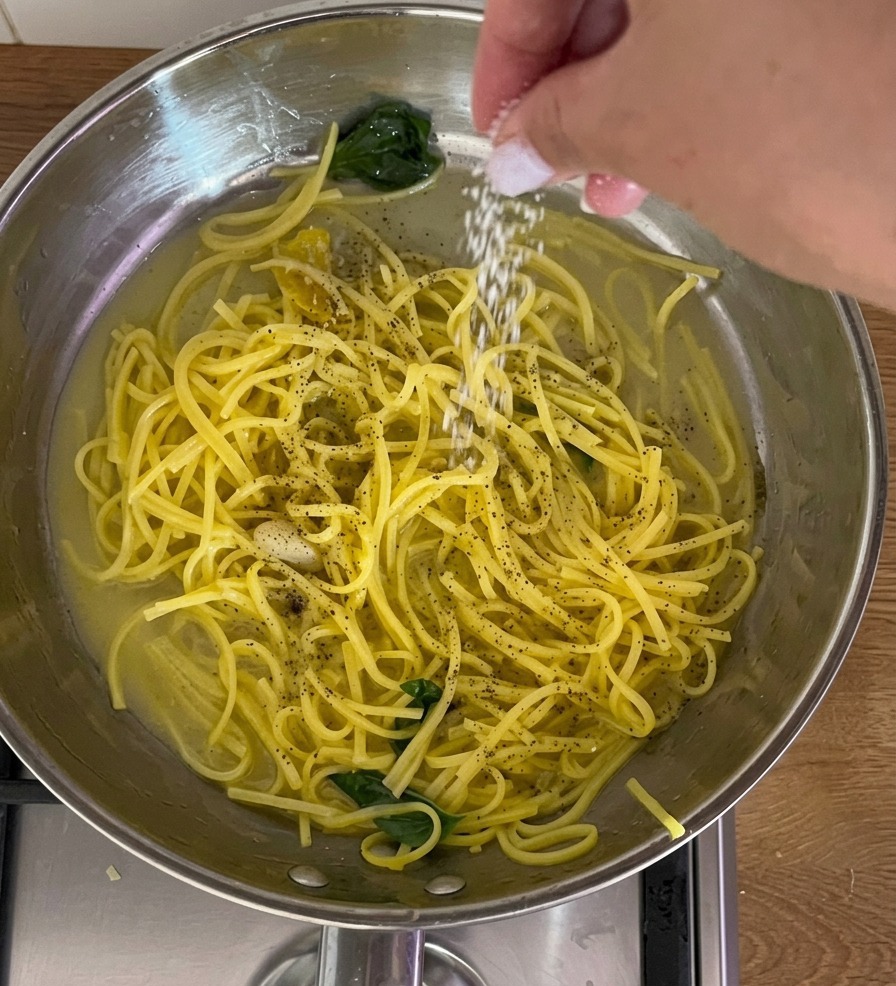 A close-up shot of a hand sprinkling fine white salt over a stainless steel pan filled with yellow spaghetti, fresh basil leaves, and cracked black pepper in a light sauce.