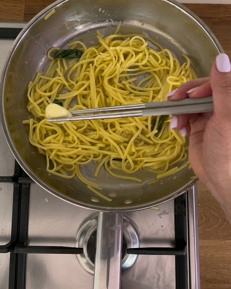 Metal chopsticks picking a softened garlic clove out of a stainless steel pan filled with yellow pasta and basil.